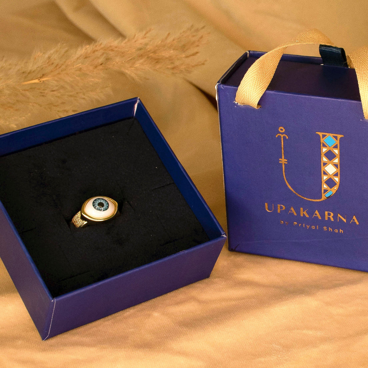 Gold-toned evil-eye ring featuring a round white enamel face with a blue and black eye motif, accented by a textured gold band. Displayed in an open navy blue Upakarna jewellery box with a black velvet insert and a matching navy gift bag with gold branding. Warm beige satin styling and soft golden lighting create a premium luxury gifting aesthetic.