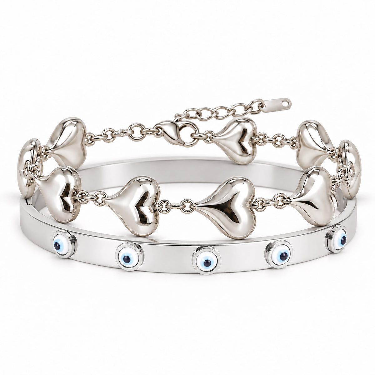 A product photo of a stacked set of silver-tone bracelets featuring a linked chain with smooth puffy heart charms and a solid bangle decorated with blue-and-white evil eye motifs.