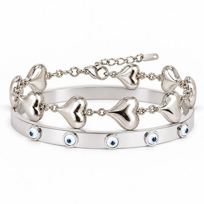A product photo of a stacked set of silver-tone bracelets featuring a linked chain with smooth puffy heart charms and a solid bangle decorated with blue-and-white evil eye motifs.