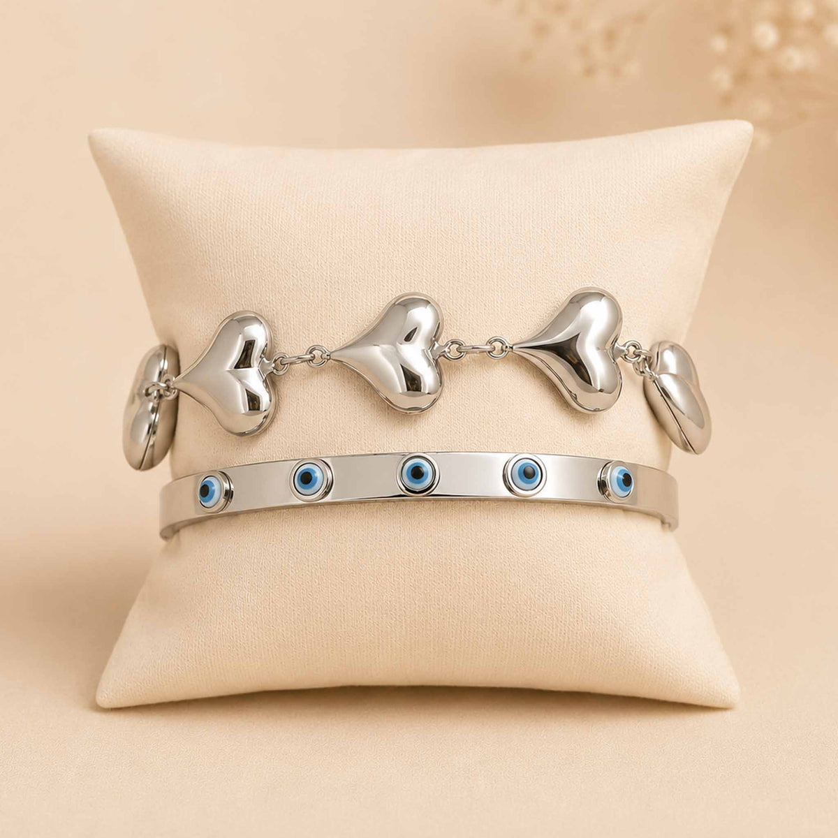 Silver bracelet stack displayed on a cream cushion, featuring a chunky heart-link bracelet and an enamel Evil Eye bangle.