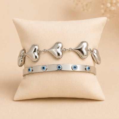 Silver bracelet stack displayed on a cream cushion, featuring a chunky heart-link bracelet and an enamel Evil Eye bangle.