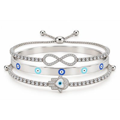 A product photo of a stacked set of silver-tone bracelets featuring an adjustable crystal band with an infinity symbol, a solid bangle with blue-and-white evil eye motifs, and a crystal tennis bracelet centered with a Hamsa hand charm containing a heart-shaped evil eye.