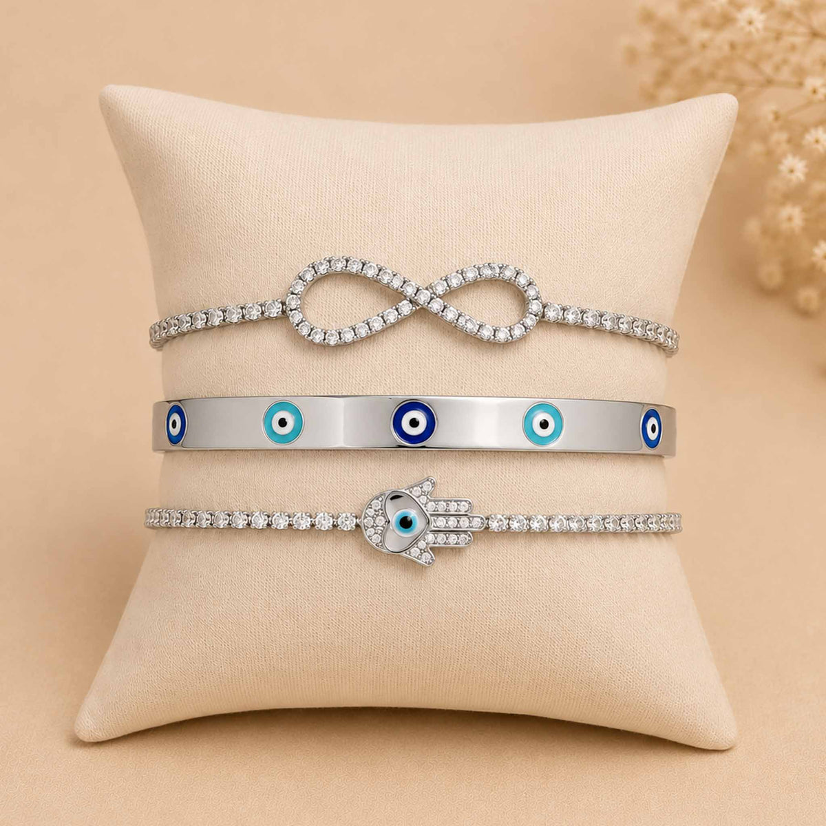 Silver bracelet stack displayed on a cream cushion, featuring an infinity crystal bracelet, enamel Evil Eye bangle, and sparkling Hamsa bracelet.