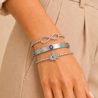 Silver bracelet stack worn on a woman’s wrist, featuring an infinity crystal bracelet, enamel Evil Eye bangle, and sparkling Hamsa bracelet.