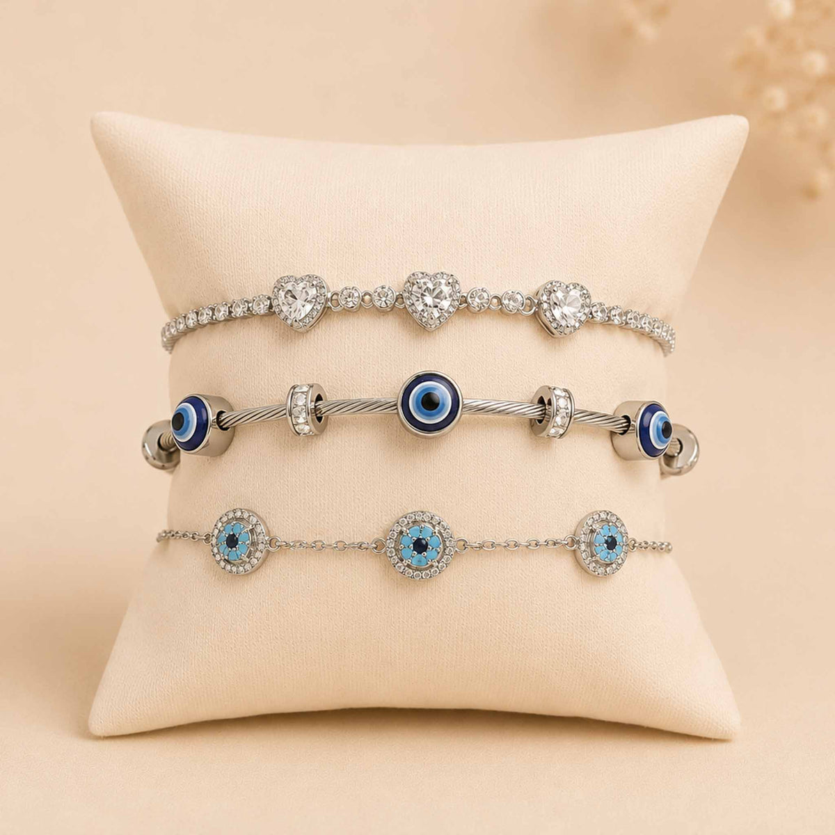Silver bracelet stack displayed on a cream cushion, featuring a sparkling triple-heart bracelet, Evil Eye bangle, and blue Evil Eye bracelet.