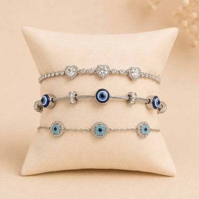 Silver bracelet stack displayed on a cream cushion, featuring a sparkling triple-heart bracelet, Evil Eye bangle, and blue Evil Eye bracelet.