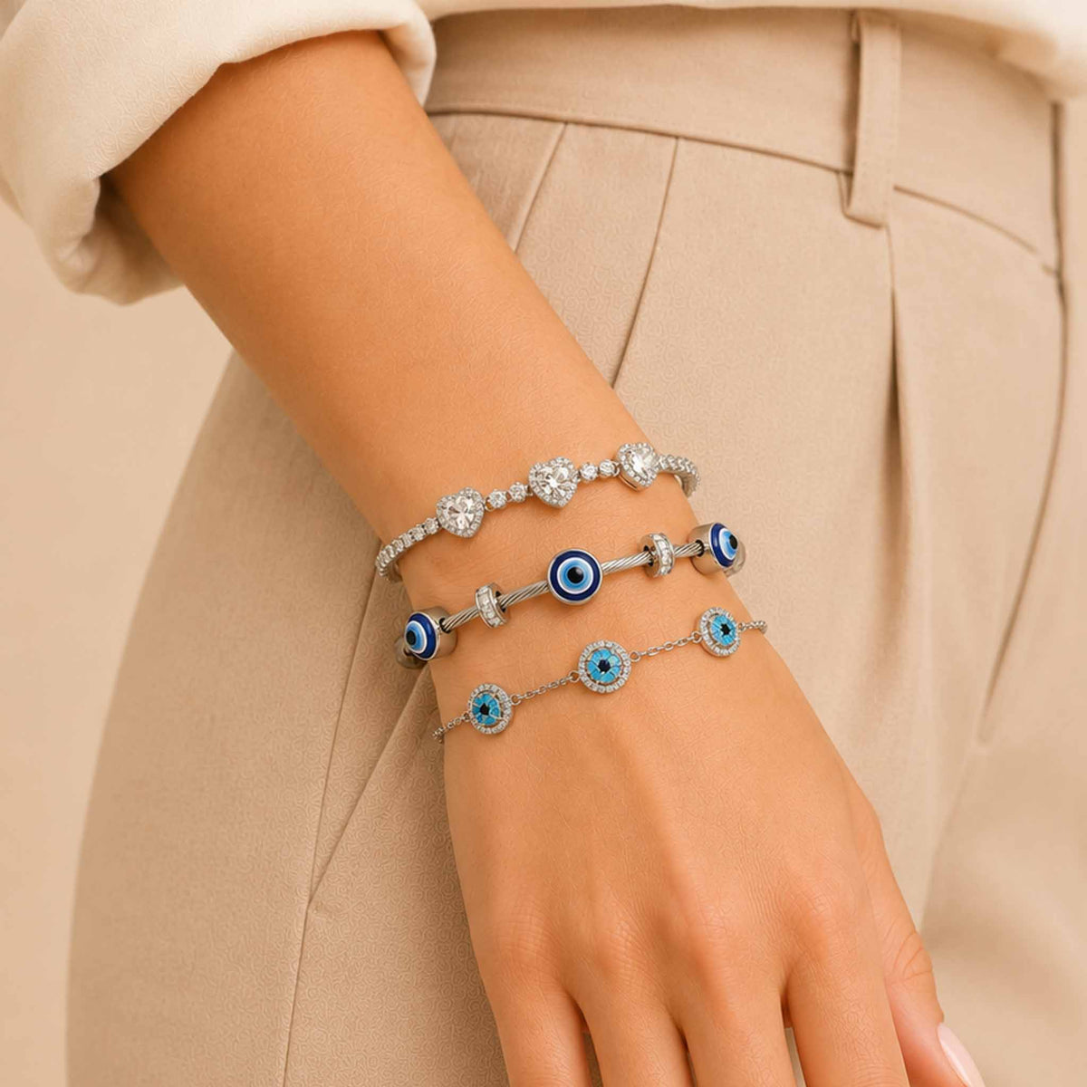 Silver bracelet stack worn on a woman’s wrist, featuring a sparkling triple-heart bracelet, Evil Eye bangle, and blue Evil Eye bracelet.