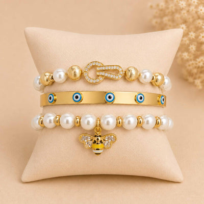 Golden jewellery stack displayed on a cream cushion, featuring a pearl bracelet with crystal clasp, Evil Eye cuff bangle, and pearl bracelet with a bee charm.