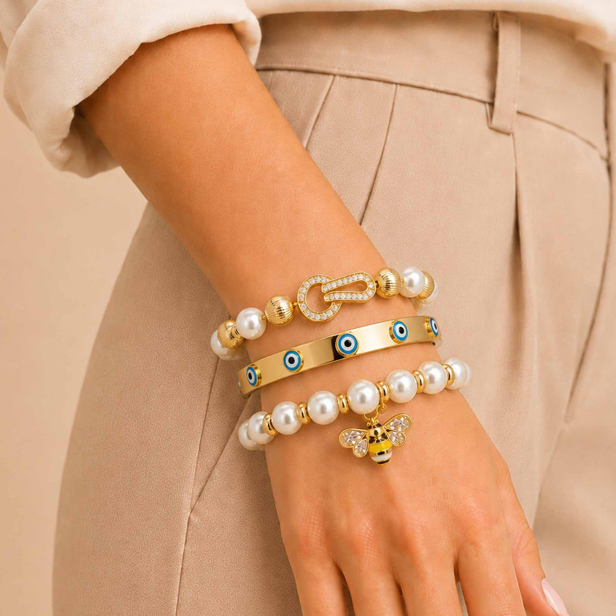 Golden pearl and Evil Eye bracelet stack styled on a woman's wrist with a neutral outfit, featuring a bee charm and sparkling clasp detail.