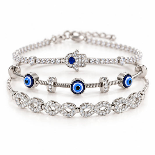 A product photo of a stacked set of silver-tone bracelets featuring a crystal-paved Hamsa hand with a blue stone, a twisted cable wire band with blue-and-white evil eye charms, and a linked infinity symbol bracelet covered in rhinestones.