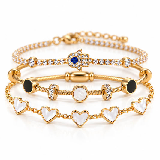 A product photo of a stacked set of gold bracelets featuring a crystal tennis chain with a Hamsa hand charm, a cable wire band with black and white stone stations, and a chain link bracelet with white mother-of-pearl heart charms.