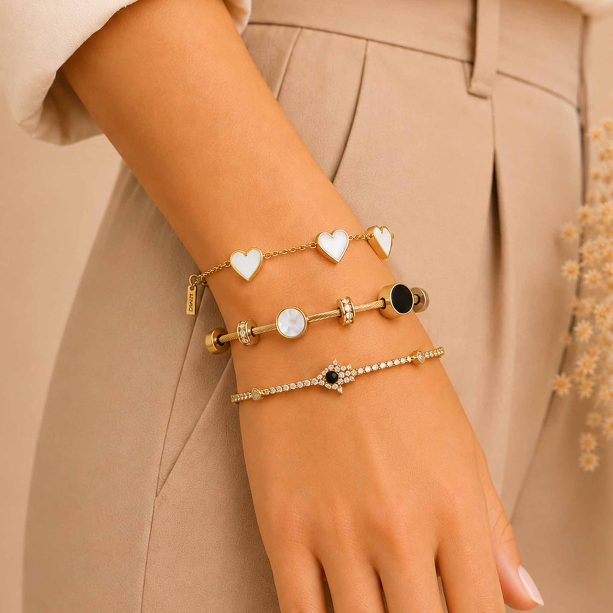 Gold-tone bracelet stack worn on a woman’s wrist, featuring white heart charms, a textured bangle with mother-of-pearl and black accents, and a sparkling Evil Eye tennis bracelet.