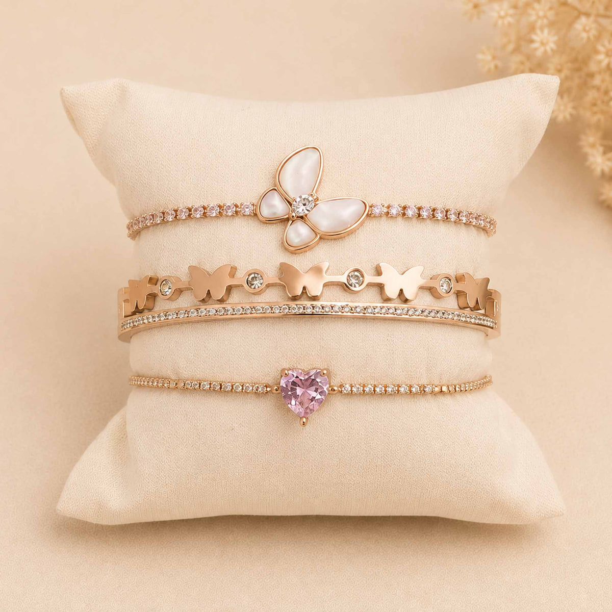 Rosegold bracelet stack displayed on a cream cushion, featuring a mother of pearl butterfly bracelet, butterfly bangle, and sparkling pink heart bracelet.