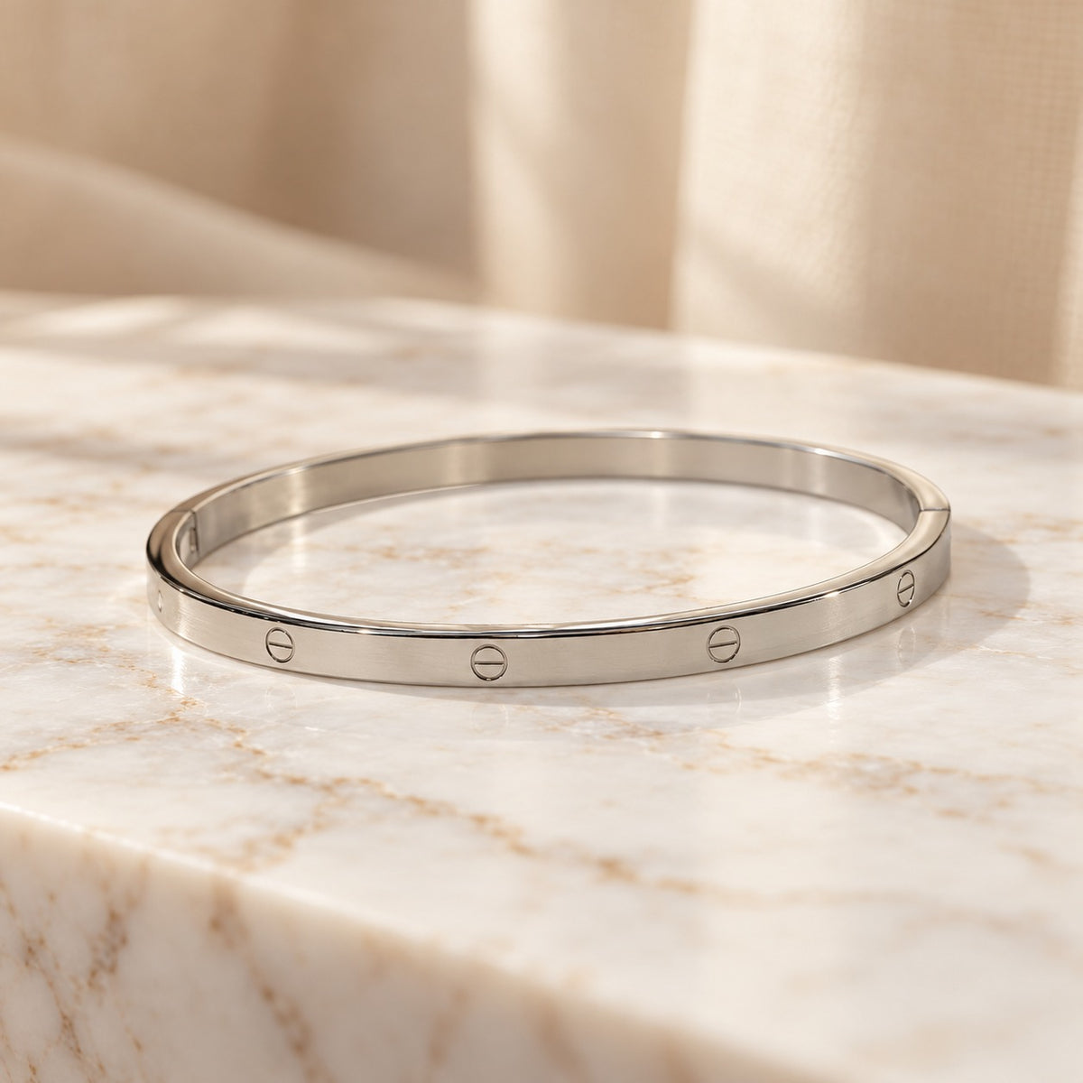 Silver-tone minimalist bangle displayed on a beige marble surface, featuring a sleek polished finish and three engraved screw-inspired details, styled against a soft cream fabric backdrop for an elegant, premium jewellery product photograph.