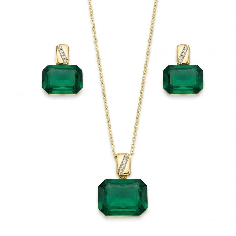 A gold jewelry set isolated on a plain white background, featuring a fine-chain necklace with a large emerald-green, emerald-cut gemstone pendant suspended from a polished gold bail with a diagonal pave-set crystal accent. The set is paired with matching stud earrings that replicate the same emerald-cut green stone and pave-accented gold bail design for a sophisticated finish.