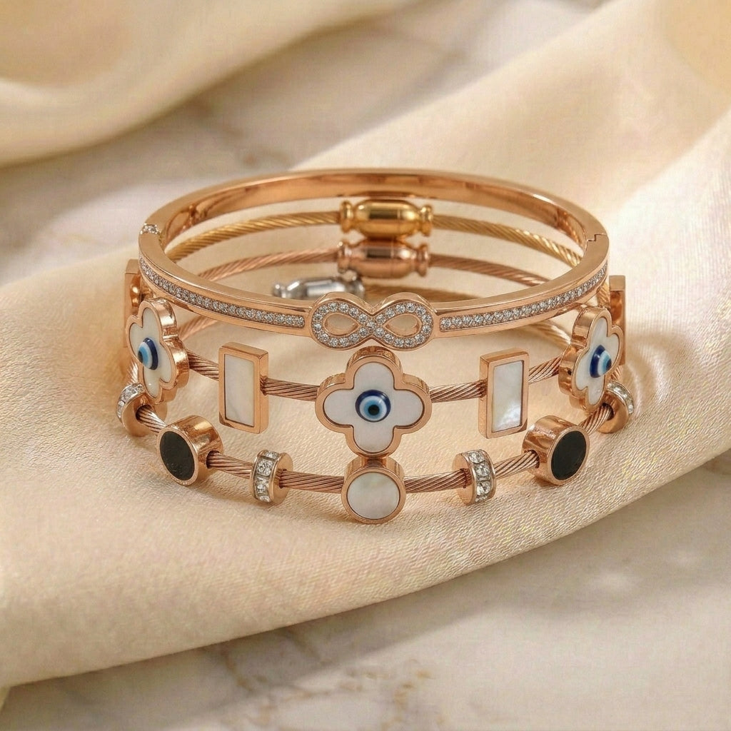 Rose gold anti-tarnish bracelet stack featuring a crystal infinity bangle, white clover and mother-of-pearl charms, blue evil eye accents, and sparkling crystal details, elegantly displayed on soft satin fabric.