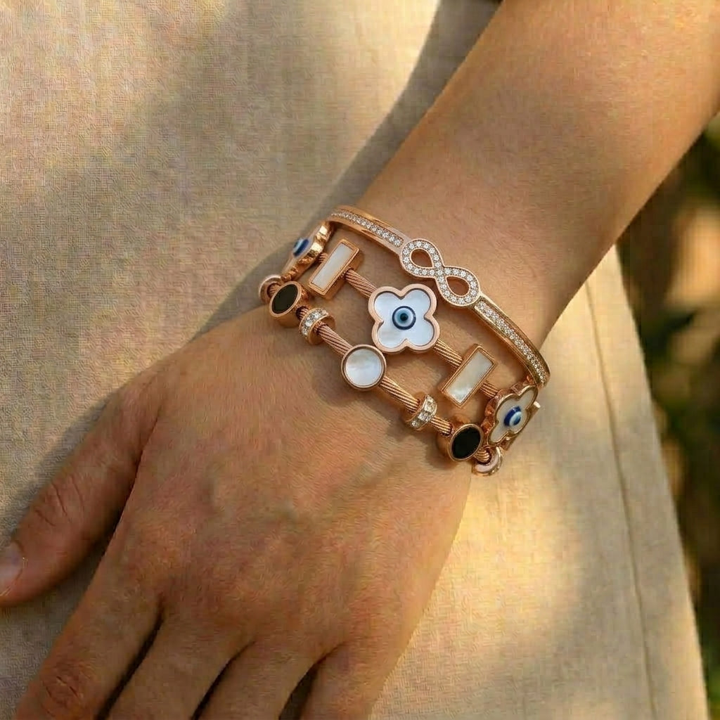 Rose gold anti-tarnish bracelet stack featuring a crystal infinity bangle, white clover and mother-of-pearl charms, blue evil eye accents, and sparkling crystal details, elegantly worn on the wrist in natural sunlight.
