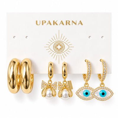 Three pairs of gold-plated statement earrings displayed on an Upakarna jewellery card, featuring chunky tube hoops, pearl bow drop earrings with crystal accents, and sparkling evil eye drop hoops, arranged on a clean white background with a premium polished finish.