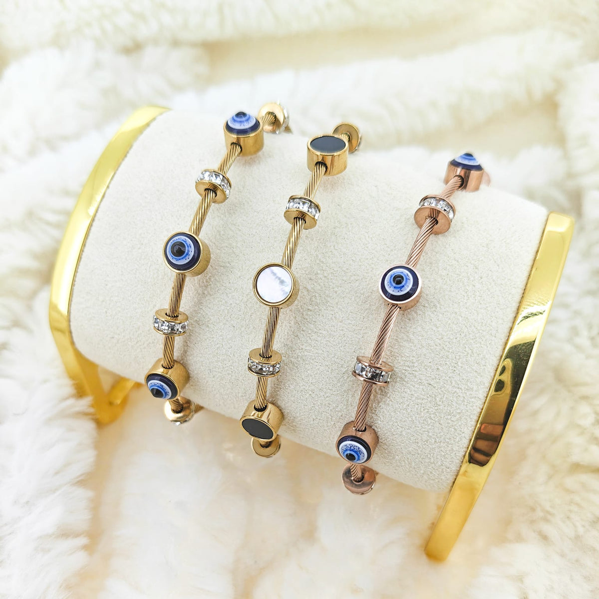 A studio product photograph of a curated trio of mixed-metal cable-wire bracelets displayed on a white velvet cushion against a fluffy white background. The set features gold-toned and rose-gold-toned wire bands accented with alternating dark blue and white evil eye charms and sparkling crystal station spacers, alongside a central gold-toned wire bracelet featuring alternating black and white circular inlay charms.   
