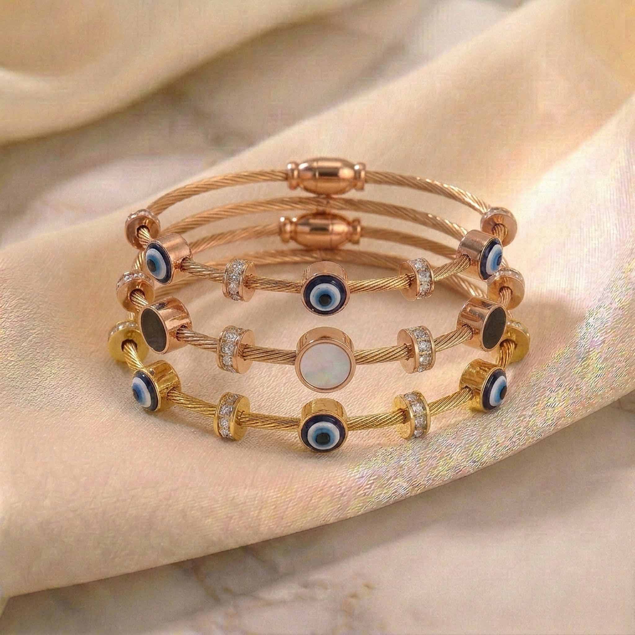 Luxury gold and rose gold anti-tarnish bracelet stack featuring blue evil eye charms, crystal-studded cable bracelets, and a mother-of-pearl center charm, displayed on soft satin fabric.