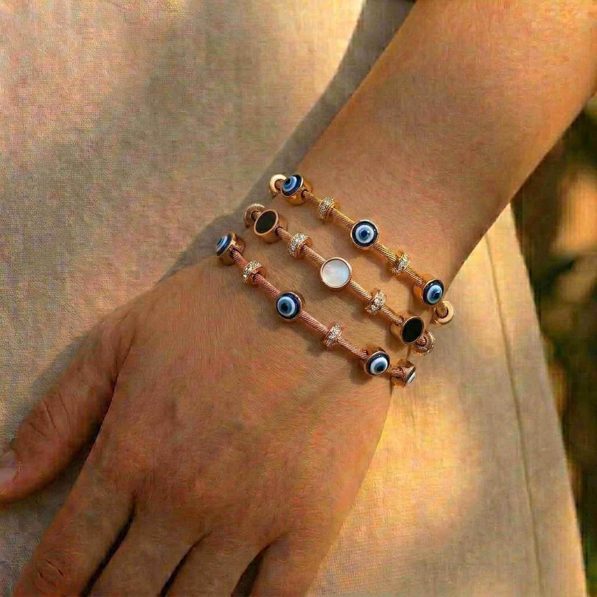 Gold and rose gold anti-tarnish bracelet stack featuring blue evil eye charms, crystal-studded cable bracelets, and a mother-of-pearl center charm, elegantly worn on the wrist in natural sunlight.