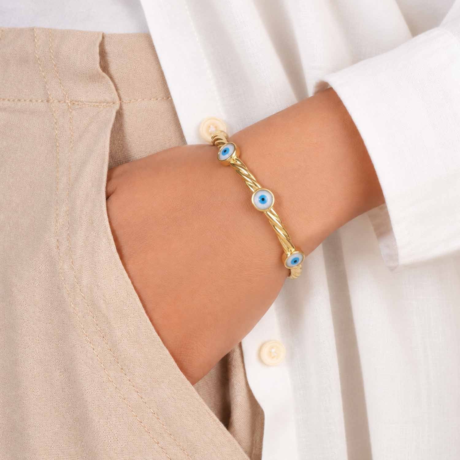 Gold twisted cuff bangle featuring three blue evil eye motifs, worn on the wrist with a neutral beige and white outfit.