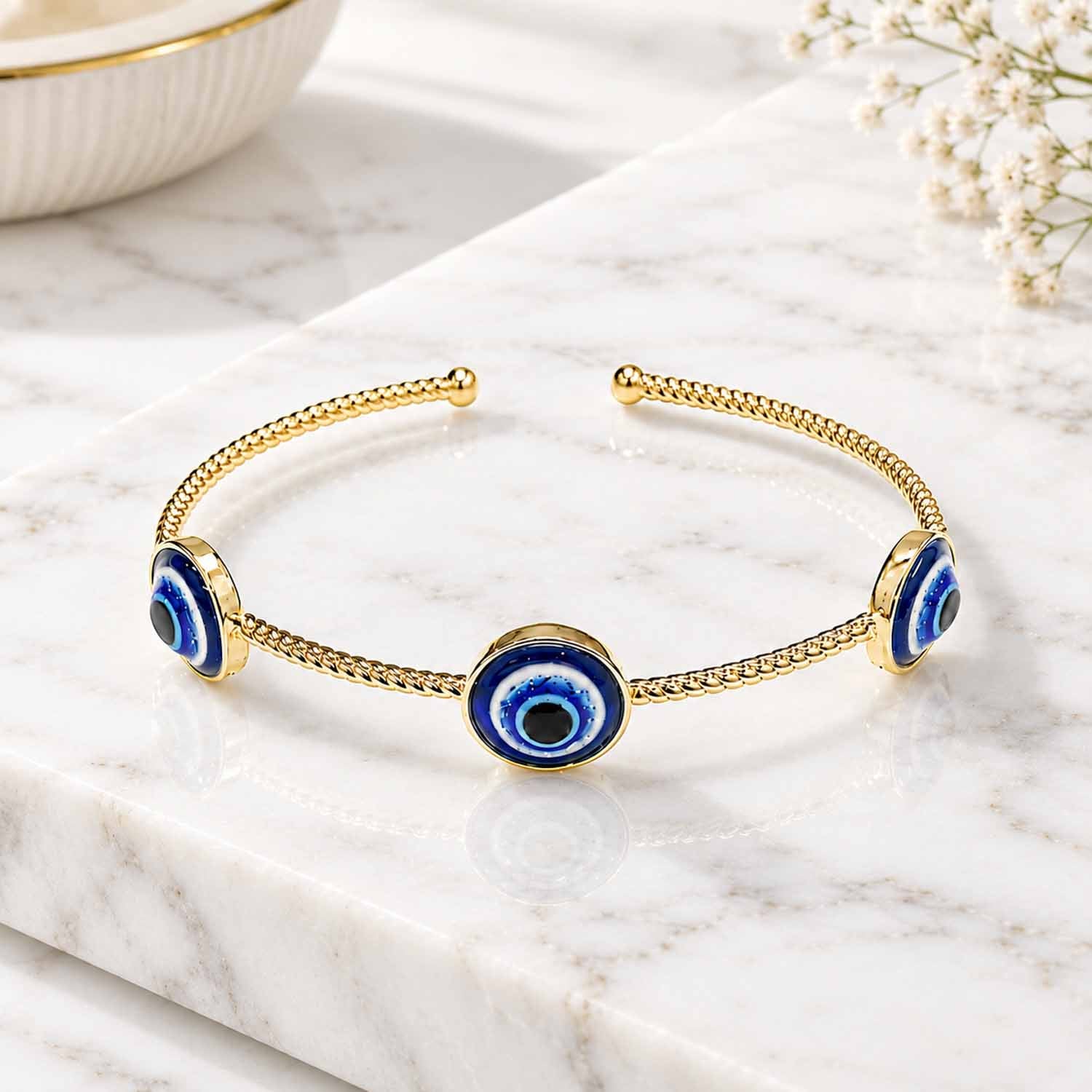 Gold evil eye cuff bangle with three blue enamel evil eye charms, displayed on a white marble surface with soft natural lighting and subtle reflections.