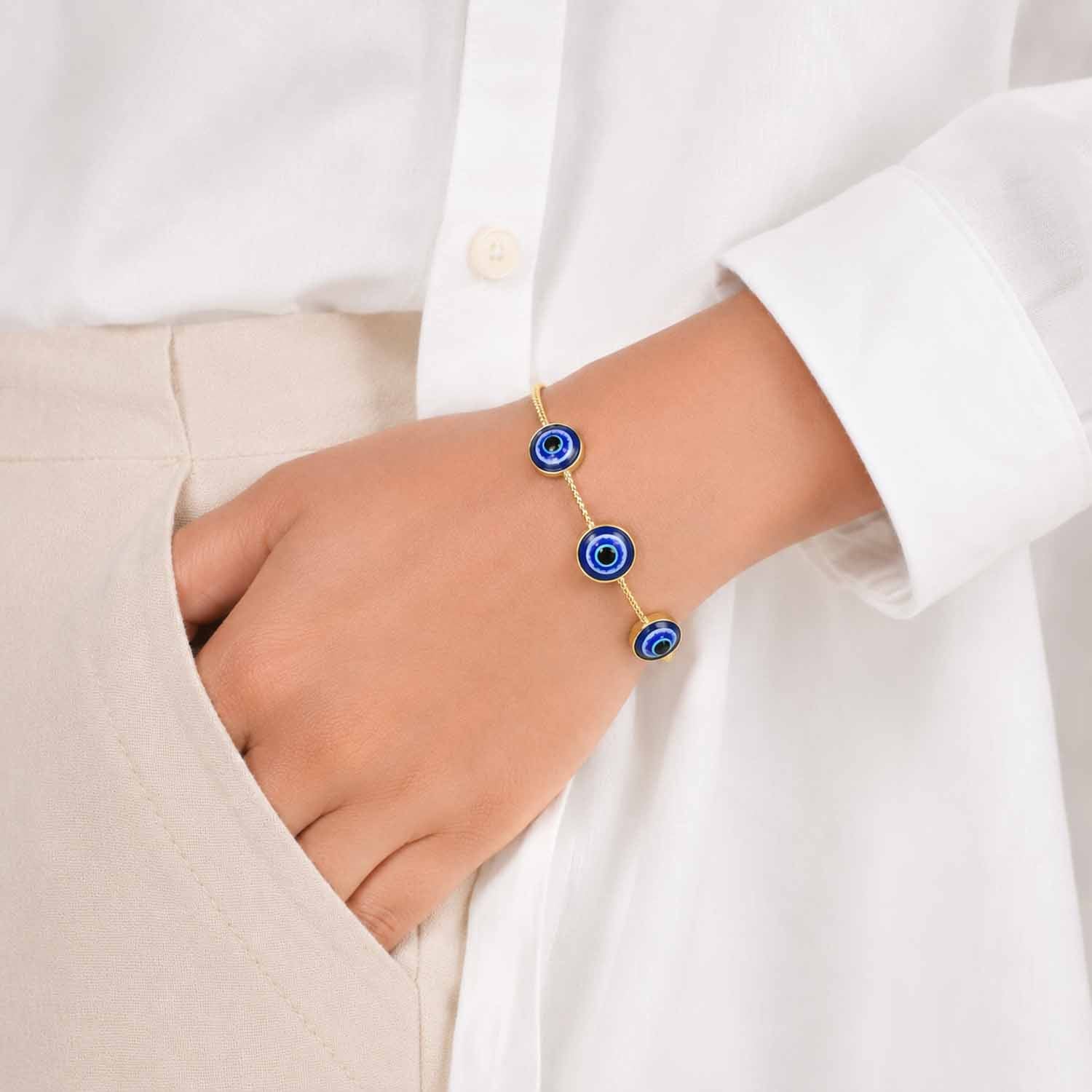 Delicate gold cuff bangle with blue evil eye charms, worn on the wrist with a neutral white and beige outfit.