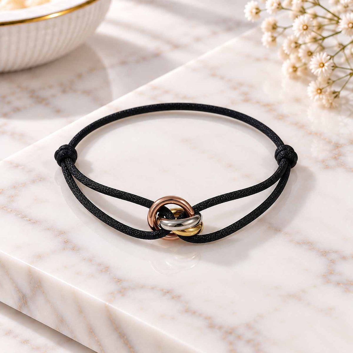 Three-tone intertwined ring charm bracelet with an adjustable black cord, displayed on a marble surface.