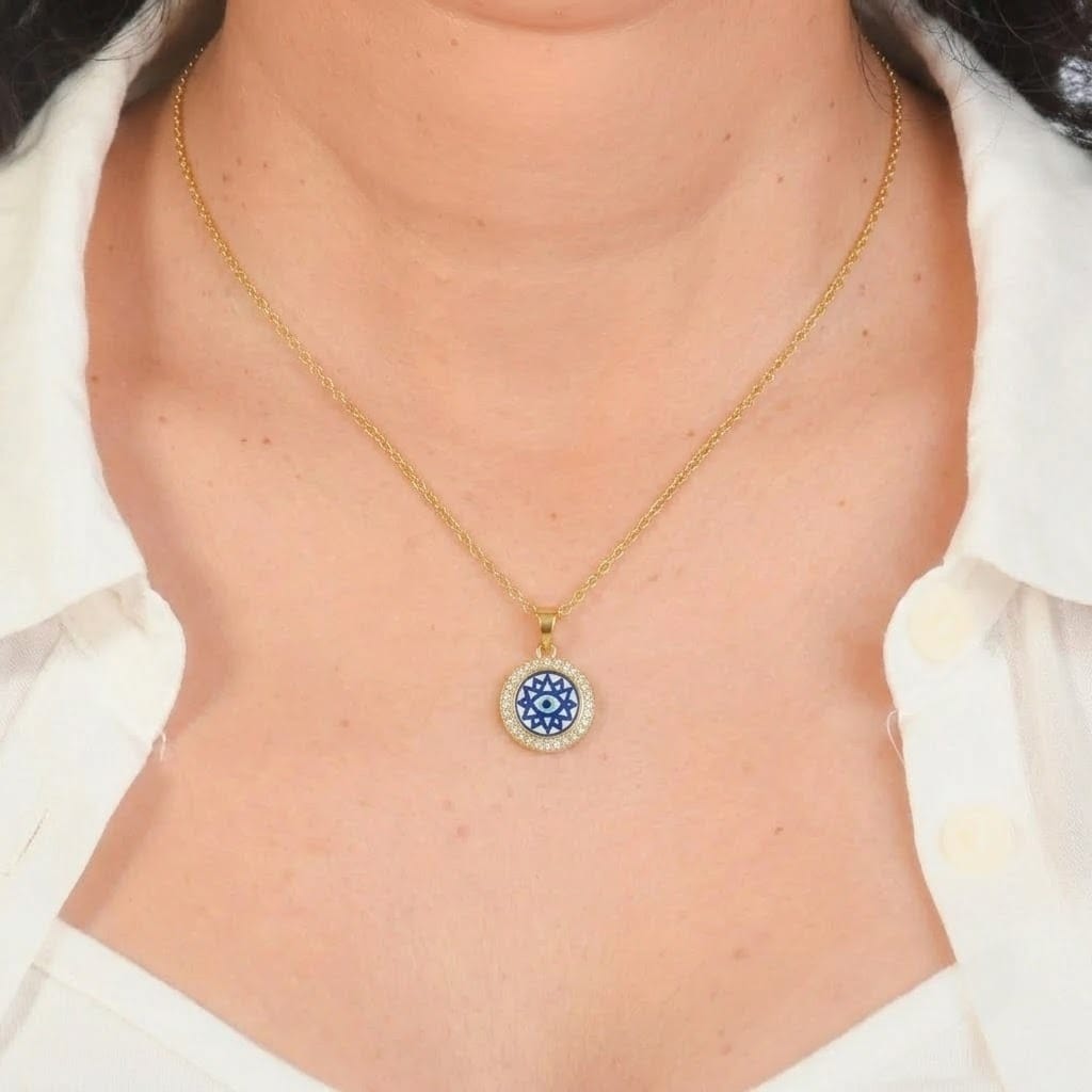 Close-up view of a model wearing a fine gold chain necklace with a circular pendant. The pendant features a pavé ring of sparkling clear crystals encircling a navy blue and white star-like geometric Evil Eye center, resting on the chest framed by an open white collared shirt set against a dark gray border.