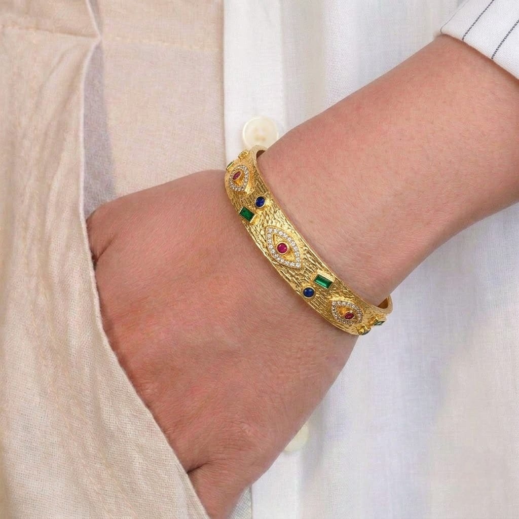 A close-up of a person’s wrist wearing a gold-toned, intricately textured bangle featuring embedded multicolored stones, including red, blue, and green accents. The bracelet has an ornate, slightly vintage design and is styled against a soft beige outfit.