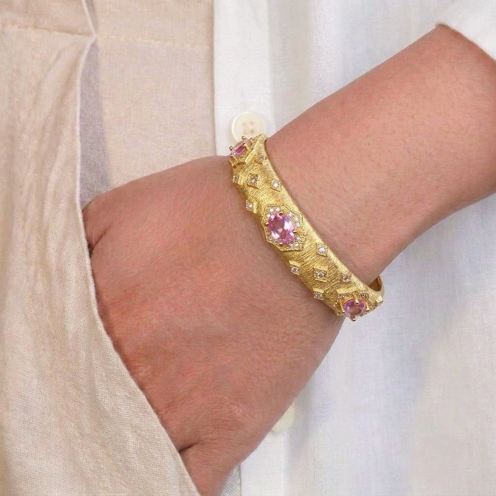 A close-up of a person’s wrist wearing a textured gold-toned bangle adorned with pink oval gemstones and small sparkling stone accents. The intricate, vintage-inspired design is styled against a soft beige outfit, giving a rich and elegant look.