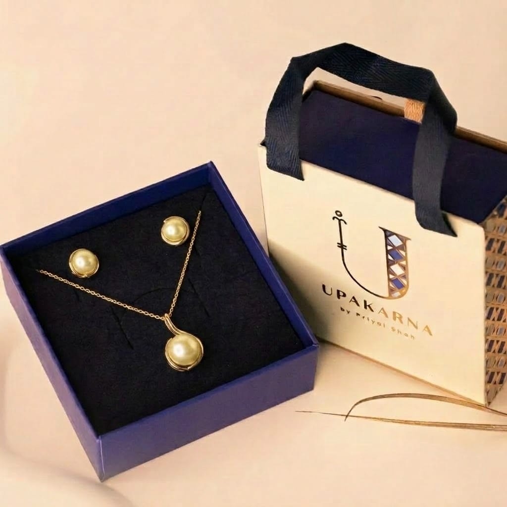 A yellow gold pearl jewelry set displayed inside a dark blue gift box alongside a cream "UPAKARNA by Priyal Shah" branded gift bag. Nestled on the navy velvet interior tray, the set features a fine-chain necklace with a pendant that houses a golden pearl cradled within a fluid, swooping curved gold setting, paired with matching stud earrings wrapped in complementary gold metal swirls.