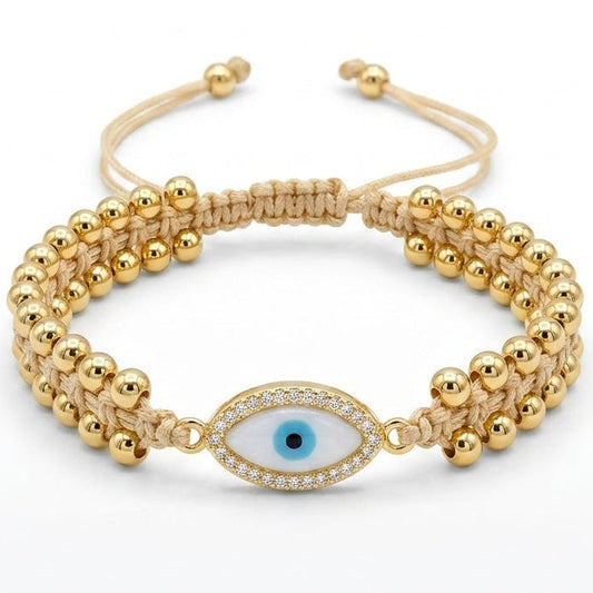  Gold-tone evil eye bracelet with polished round beads, a beige braided cord, and a blue-and-black evil eye charm bordered with sparkling clear crystals, displayed on a white background.
