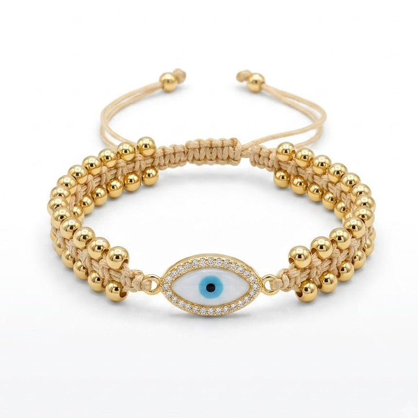  Gold-tone evil eye bracelet with polished round beads, a beige braided cord, and a blue-and-black evil eye charm bordered with sparkling clear crystals, displayed on a white background.