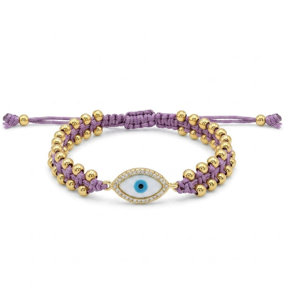 Purple braided cord bracelet featuring rows of polished gold beads and a central oval evil eye charm. The charm has a blue iris with a black center and is surrounded by sparkling clear crystals. Displayed neatly on a clean white background.