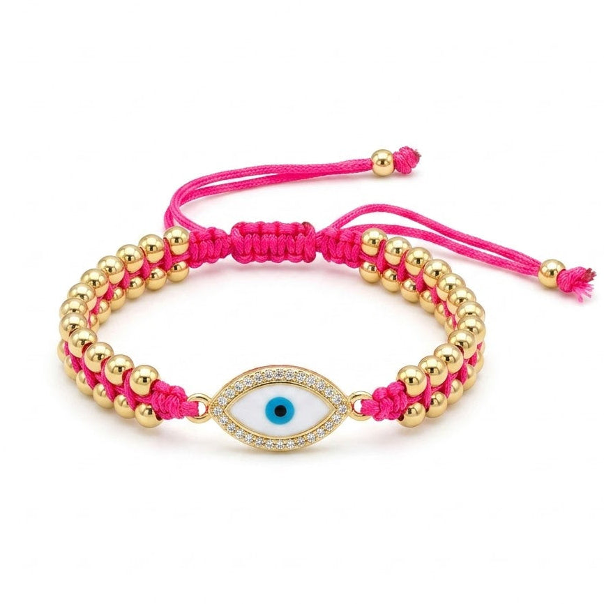 Hot pink braided cord bracelet with polished gold beads and a central oval blue evil eye charm. The charm is outlined with sparkling clear crystals and paired with adjustable pink cord ends. Displayed on a clean white background.