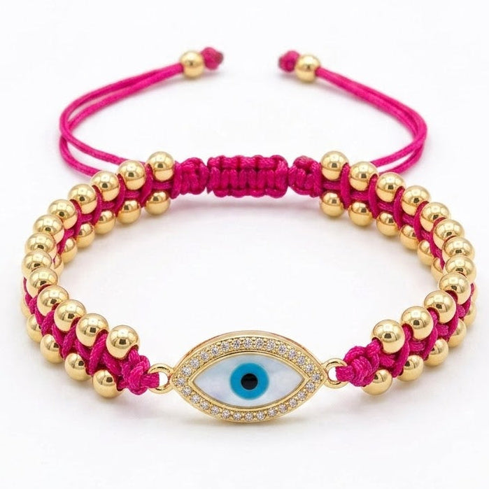 Hot pink braided cord bracelet with rows of polished gold beads and a central oval evil eye charm featuring a blue iris and black pupil. The charm is bordered with sparkling clear crystals and finished with adjustable pink cord ends. Displayed on a clean white background.