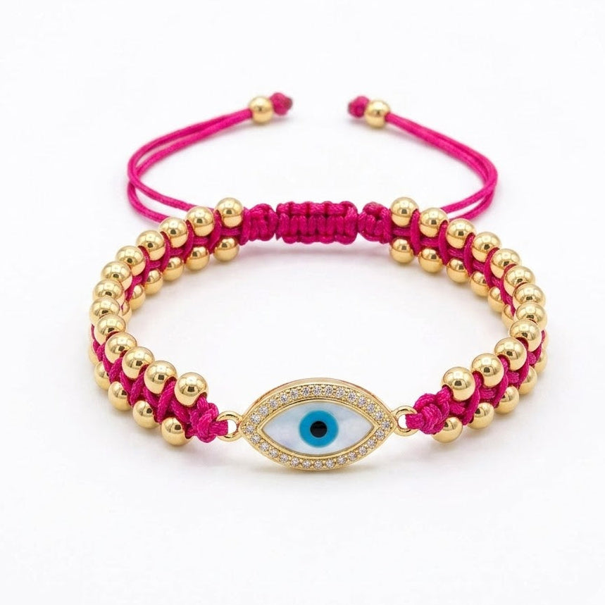 Hot pink braided cord bracelet with rows of polished gold beads and a central oval evil eye charm featuring a blue iris and black pupil. The charm is bordered with sparkling clear crystals and finished with adjustable pink cord ends. Displayed on a clean white background.