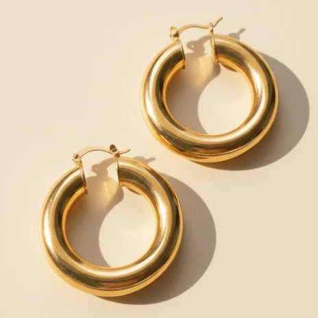 Gold-tone chunky hoop earrings with a smooth, rounded tubular design and polished finish, photographed on a warm beige background.