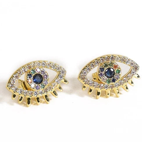 Gold-tone evil eye stud earrings featuring crystal-studded eye-shaped designs, dark blue center stones with multicolor accents, and a row of small gold-tone pointed charms along the lower edge.