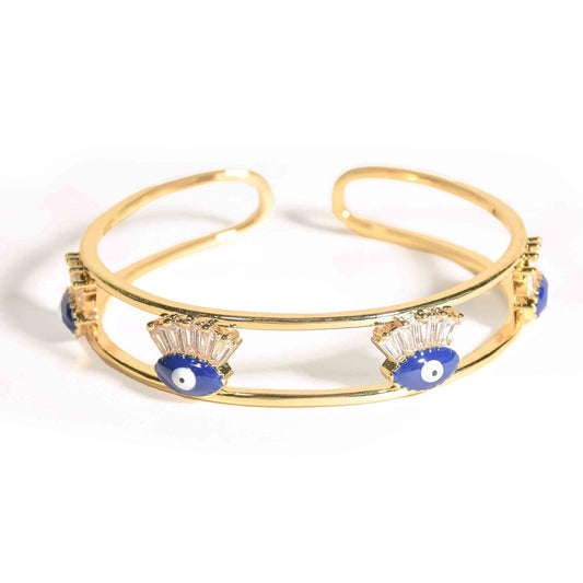 Gold-tone double-band open cuff bangle featuring four blue evil eye charms with sparkling baguette-cut crystal accents. The sleek anti-tarnish bracelet is photographed on a clean white background, showcasing its modern minimalist design and protective evil eye motif.