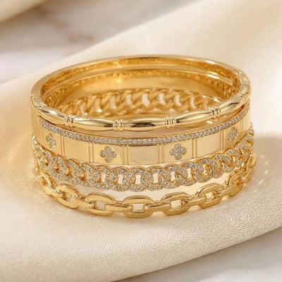 Luxury gold anti-tarnish bangle stack featuring polished chain-link bangles, crystal-studded Cuban link detailing, engraved quatrefoil motifs, and sleek gold cuffs, elegantly displayed on soft satin fabric.