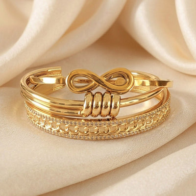 Luxury gold anti-tarnish bangle stack featuring a bold infinity centerpiece, layered open cuff design, crystal-studded chain-link detailing, and polished gold finishes, elegantly displayed on soft satin fabric.