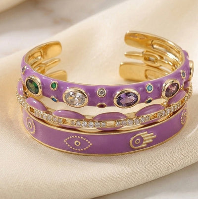 Luxury gold anti-tarnish bangle stack featuring lavender enamel bangles, colorful gemstone accents, a crystal-studded bangle, and purple evil eye motifs, elegantly displayed on soft satin fabric