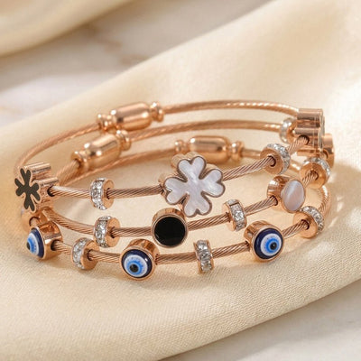 Luxury rose gold anti-tarnish bracelet stack featuring blue evil eye charms, black and white mother-of-pearl floral motifs, crystal-studded cable bracelets, and polished stainless steel detailing, elegantly displayed on soft satin fabric.