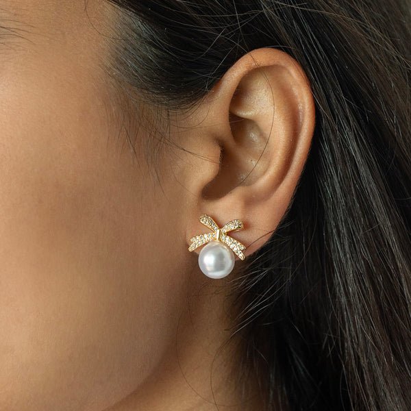 Close-up of a person wearing a gold-tone pearl drop earring with a large white pearl and a pavé crystal bow-shaped accent.