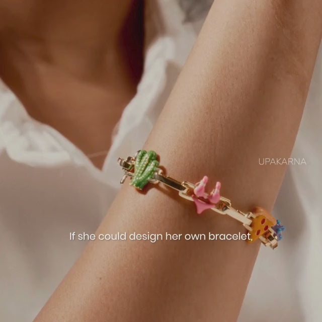 Close-up of a model wearing a gold charm bracelet with colorful cactus, pink bow, smiley face, and blue flower charms, styled on the wrist against a warm neutral background with the text.