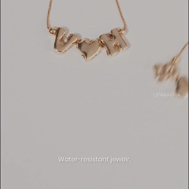 A short promotional video showcasing customizable gold jewelry pieces. It begins by displaying delicate gold chain necklaces arranged with different combinations of initial letters and heart charms. Finally, it shows two wrists wearing matching adjustable initial and heart charm bracelets side-by-side.   