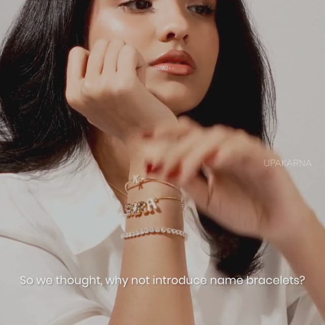 A promotional jewelry video starting with a model wearing gold accessories, transitioning to a hand with dark red nail polish sliding individual gold letter beads onto a thin chain to form the name "IRA". The video ends by showcasing several completed, dainty gold name bracelets laid out together.